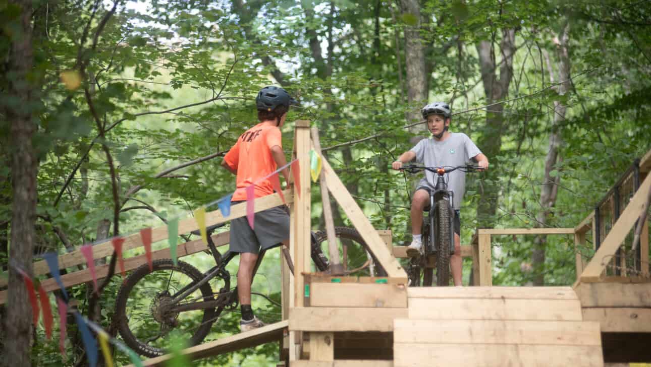 Two boys riding bikes on a wooden bridge in the woods.