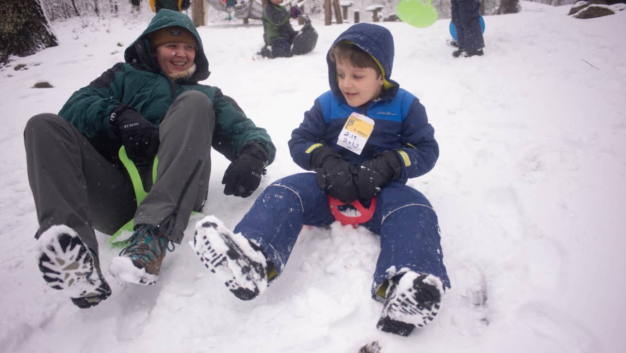 A camper and counselor sledding down a snowy hill.